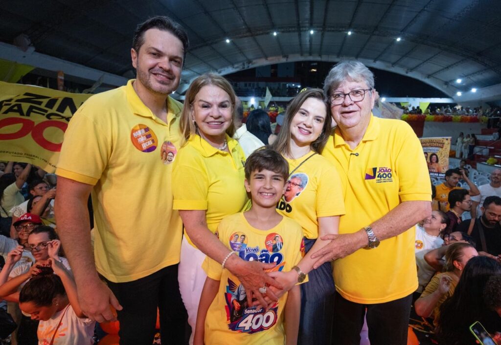 João Azevêdo posa sorrindo abraçado a familiares no palco, com o neto João Vitor à frente vestindo camiseta de apoio ao candidato.