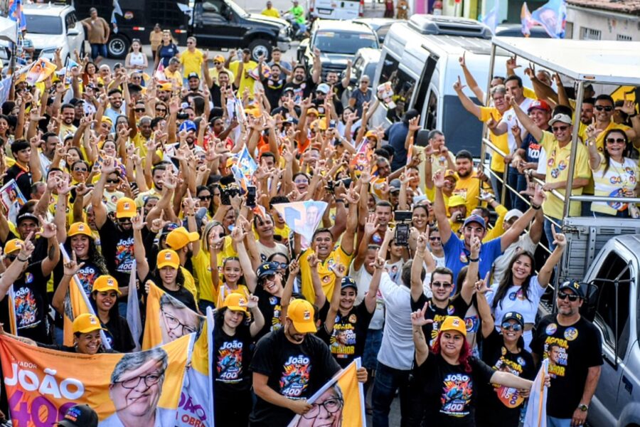 Multidão de apoiadores vestindo amarelo e segurando bandeiras com o rosto de João Azevêdo ao lado de caminhonete da comitiva