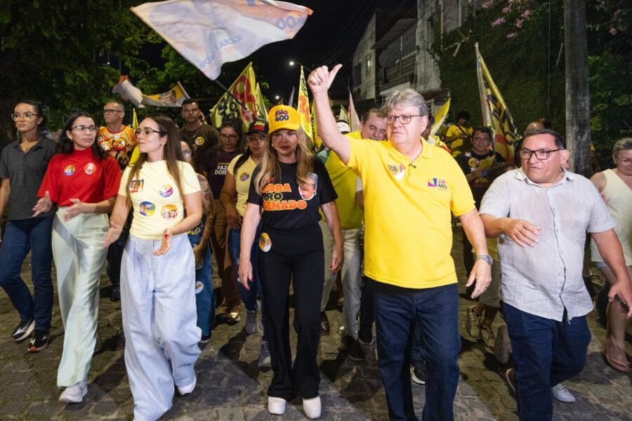 João Azevêdo caminha em rua de calçamento fazendo sinal de positivo ao lado de Camila Mariz e Ana Lins