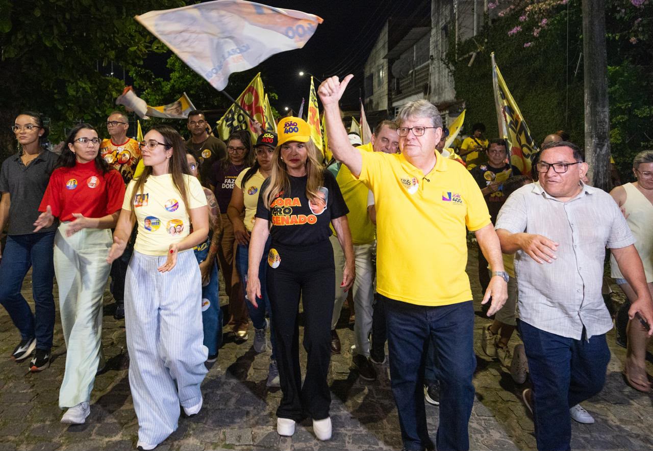 João Azevêdo caminha em rua de calçamento fazendo sinal de positivo ao lado de Camila Mariz e Ana Lins