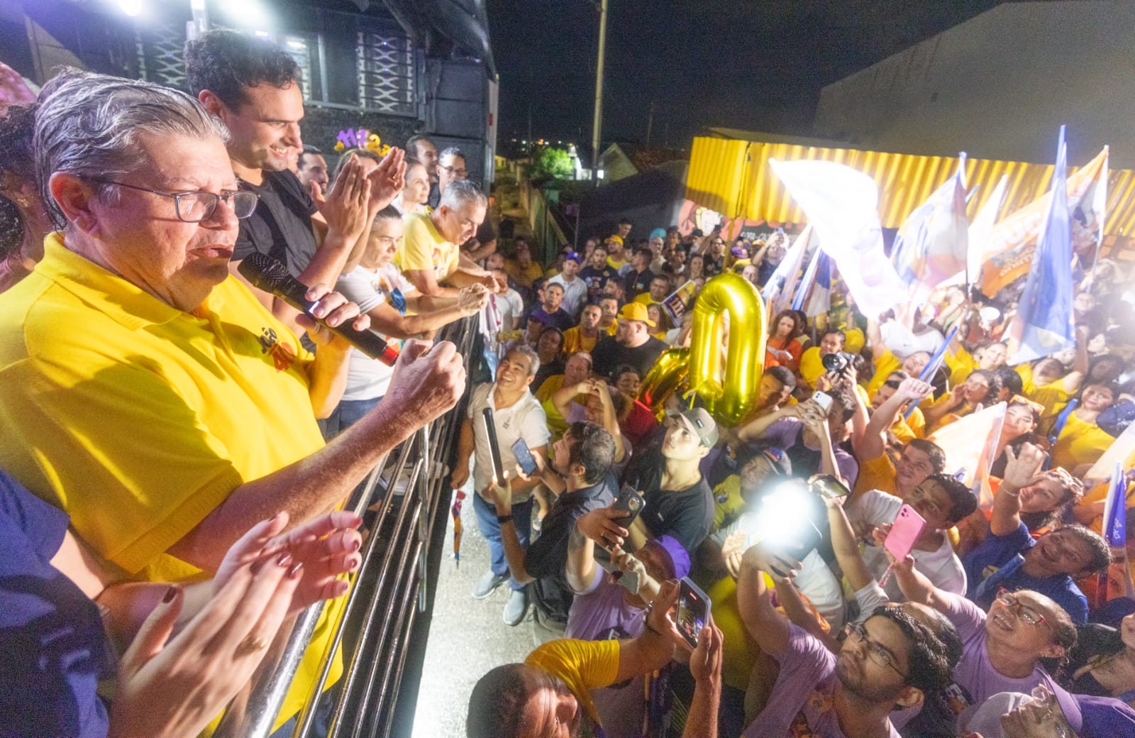 João Azevêdo discursa ao microfone ao lado de Lucas Ribeiro voltado para multidão com celulares e balão dourado.