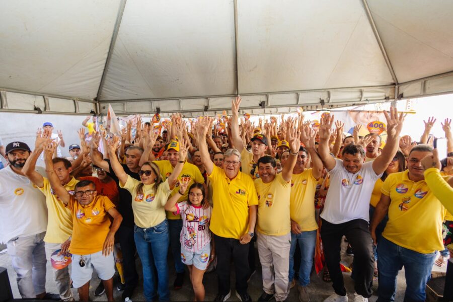 João Azevêdo posa sorrindo sob tenda com braços erguidos ao lado de uma criança e apoiadores de Areia de Baraúnas