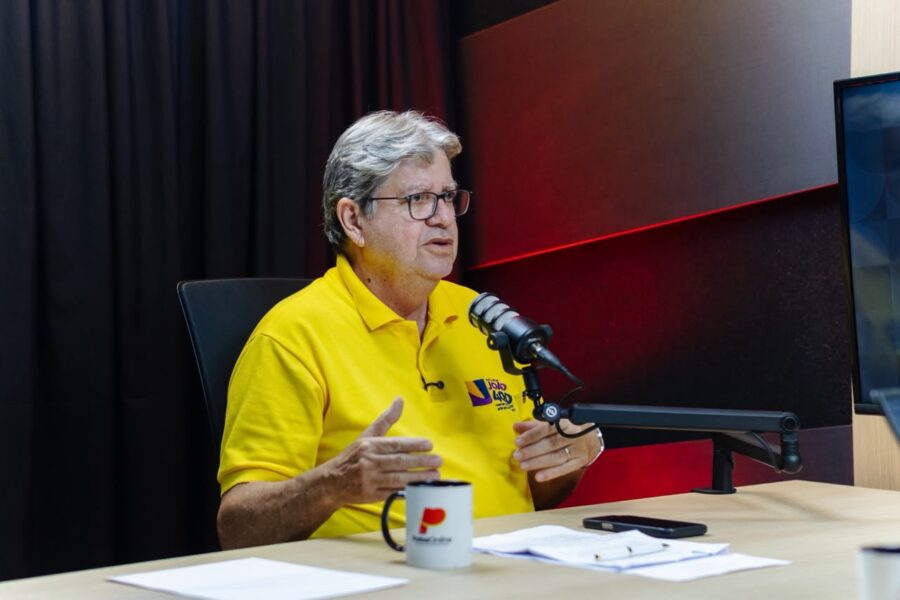 João gesticula com as mãos em frente ao microfone em estúdio de podcast e rádio.