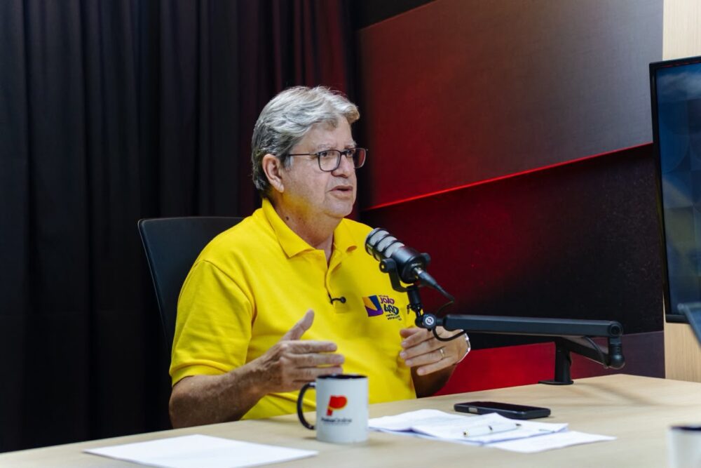 João gesticula com as mãos em frente ao microfone em estúdio de podcast e rádio.