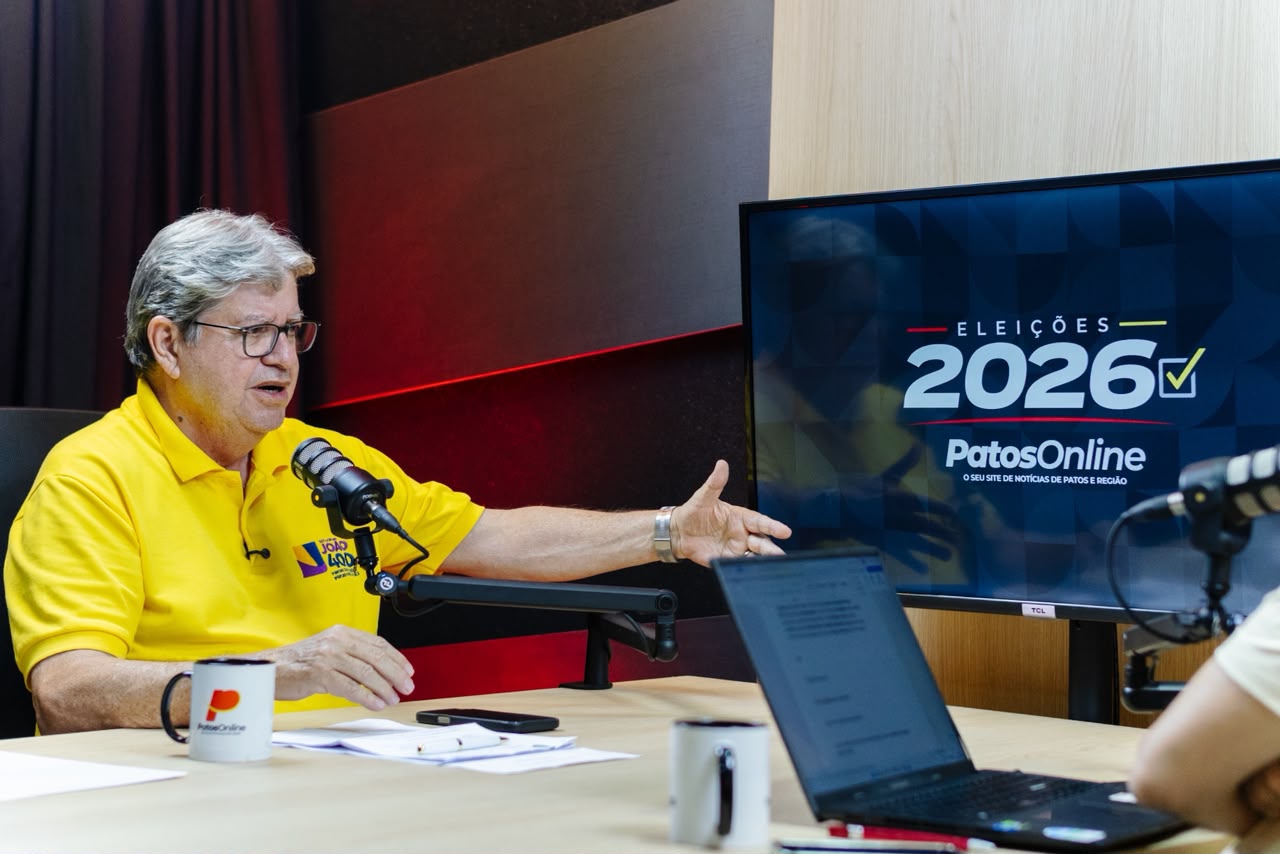 João Azevêdo de camisa polo amarela gesticula apontando para tela de monitor durante entrevista em estúdio.