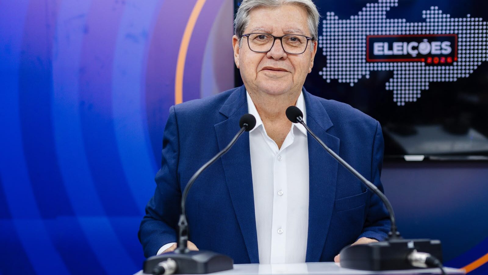 João Azevêdo de paletó azul e camisa branca fala ao microfone em púlpito de debate do Sistema Diário do Sertão.