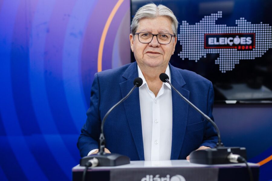 João Azevêdo de paletó azul e camisa branca fala ao microfone em púlpito de debate do Sistema Diário do Sertão.