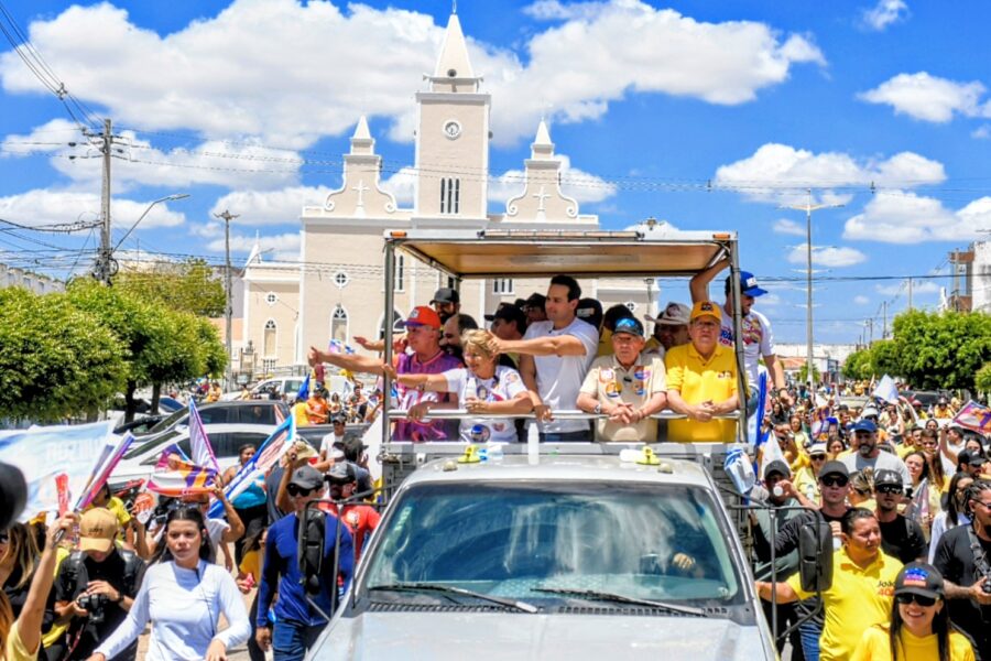 João Azevêdo de camisa polo amarela acena em veículo aberto ao lado de Lucas Ribeiro e lideranças com igreja ao fundo.