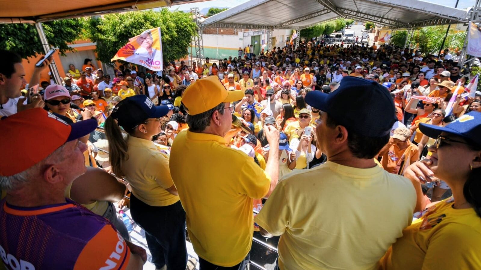 João Azevêdo de boné amarelo discursa ao microfone em palanque móvel sob tenda de frente para grande público.