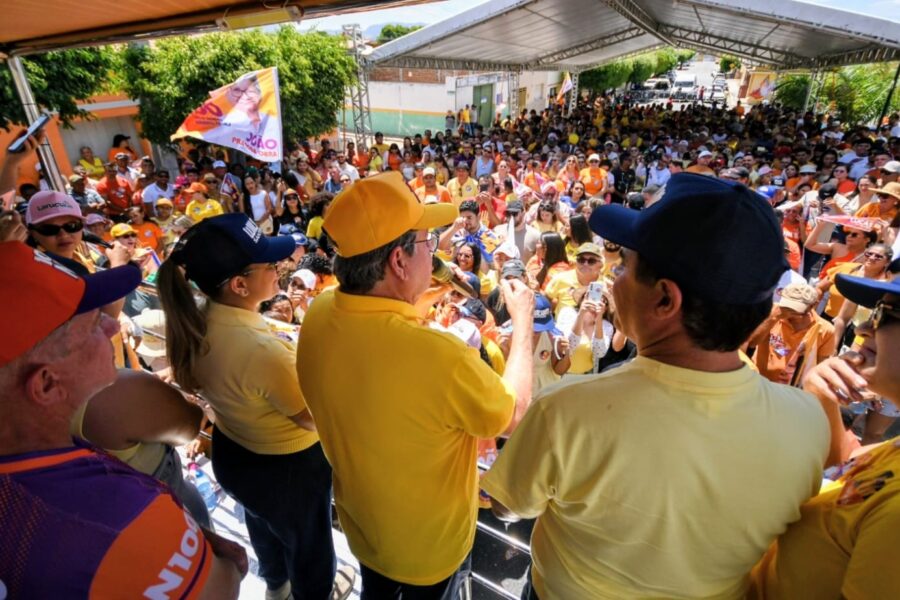 João Azevêdo de boné amarelo discursa ao microfone em palanque móvel sob tenda de frente para grande público.