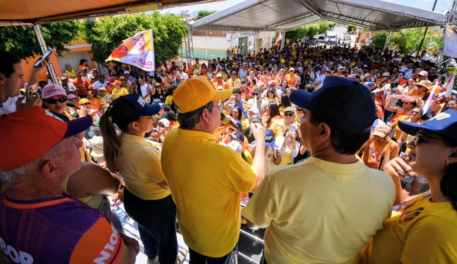 João Azevêdo de boné amarelo discursa ao microfone em palanque móvel sob tenda de frente para grande público.