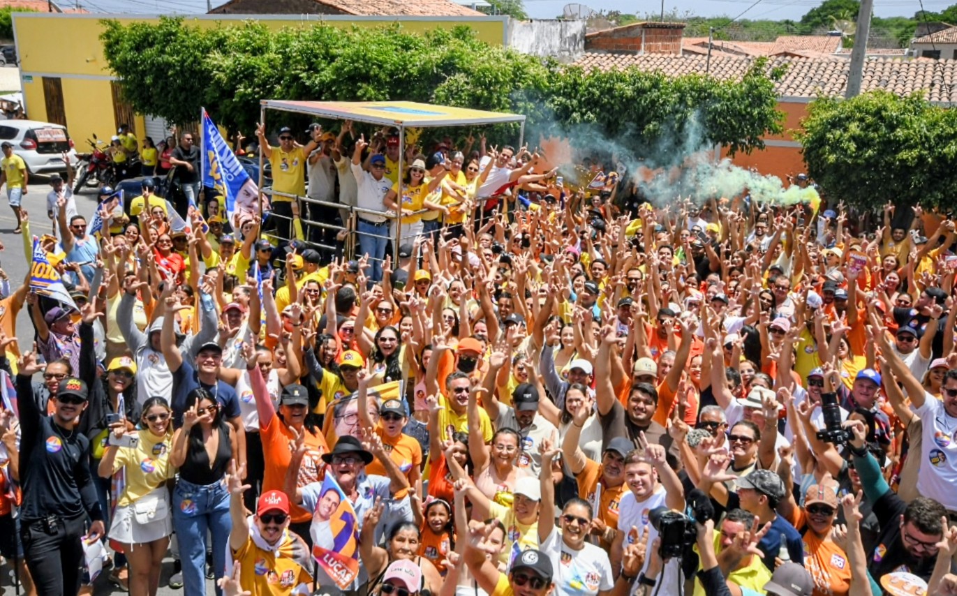 Vista ampla de grande multidão com braços levantados na rua celebrando comitiva em veículo com fumaça colorida.