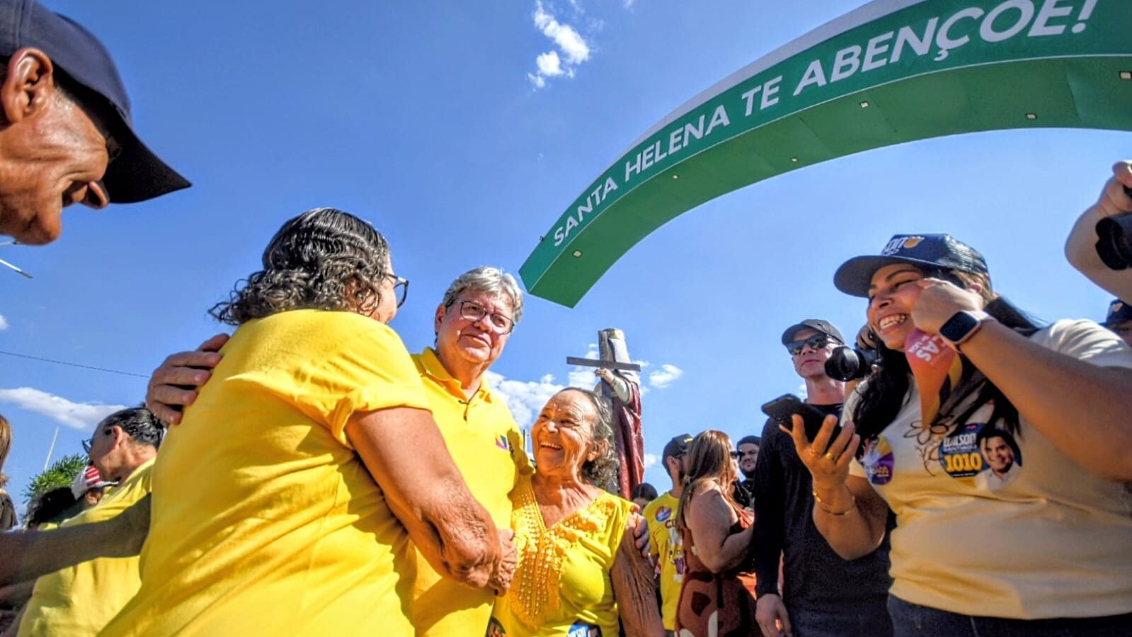 João Azevêdo abraça moradoras idosas sorridentes sob arco com a inscrição 'Santa Helena te abençoe'.