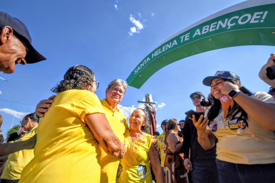 João Azevêdo abraça moradoras idosas sorridentes sob arco com a inscrição 'Santa Helena te abençoe'.