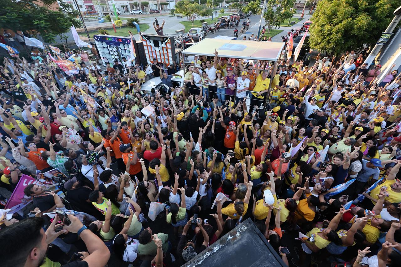 Vista superior da multidão de braços erguidos cercando o trio e a comitiva em praça de São João do Rio do Peixe.