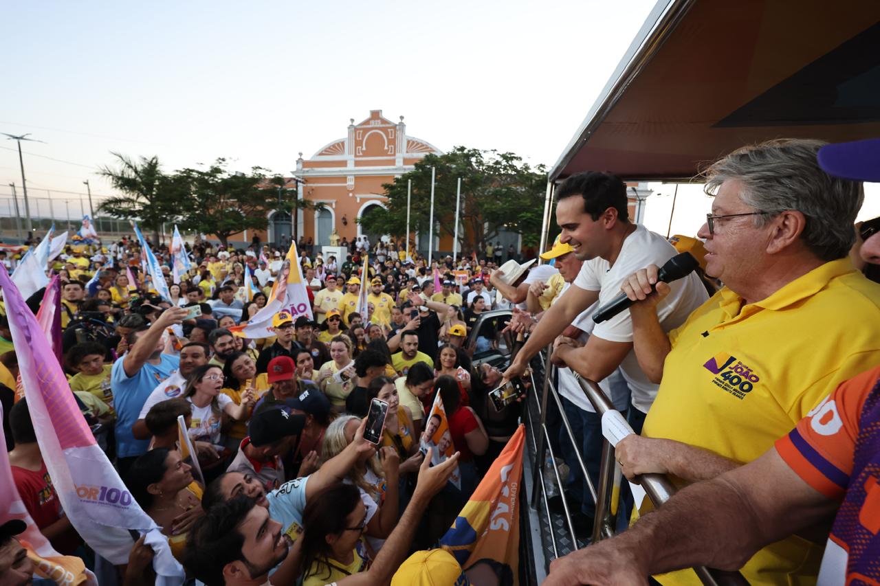 João Azevêdo de camisa amarela discursa ao microfone ao lado de Lucas Ribeiro em veículo de campanha ao entardecer.
