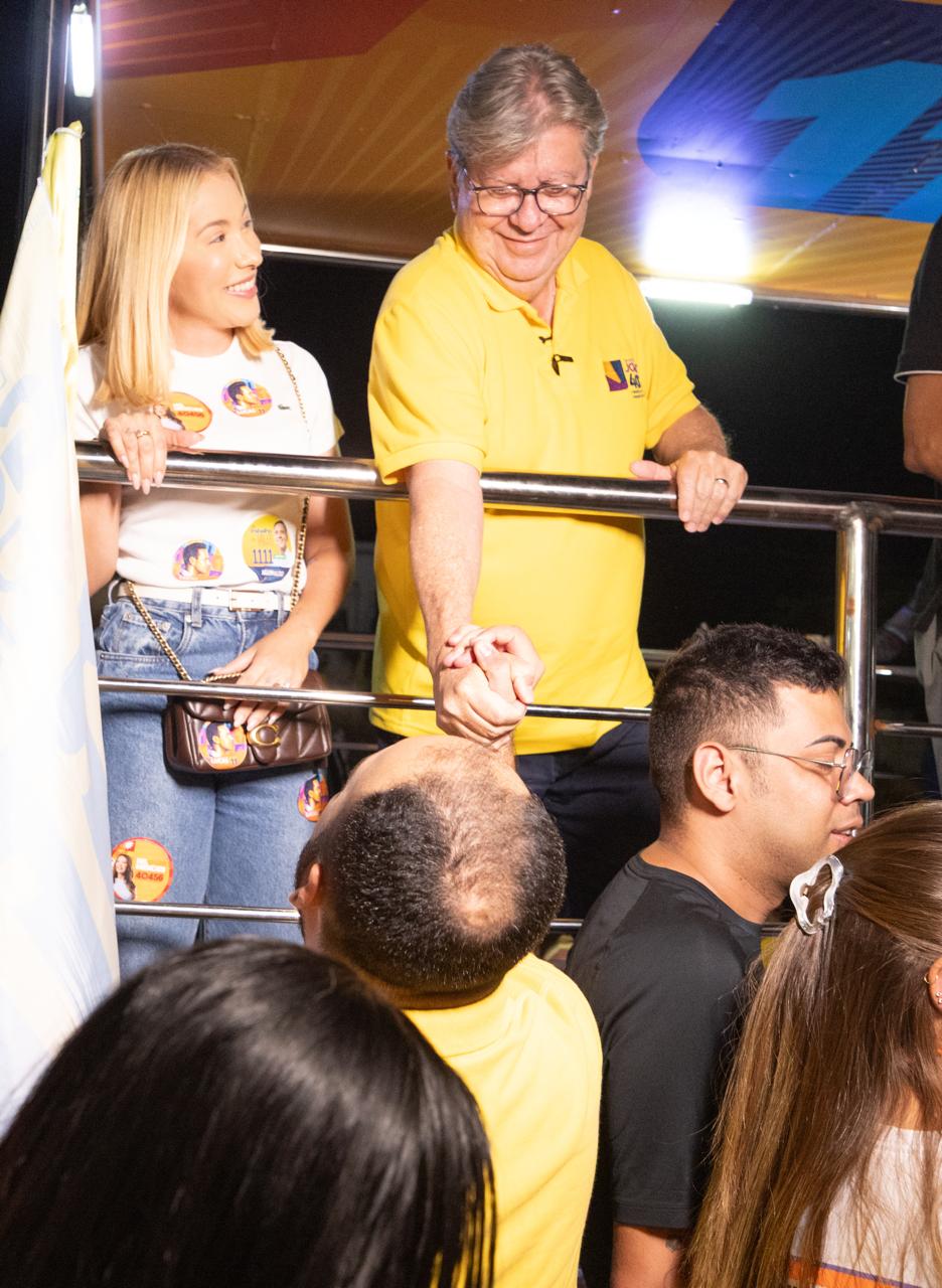 João Azevêdo sorri e segura firmemente a mão de um morador a partir do guarda-corpo de veículo de campanha.
