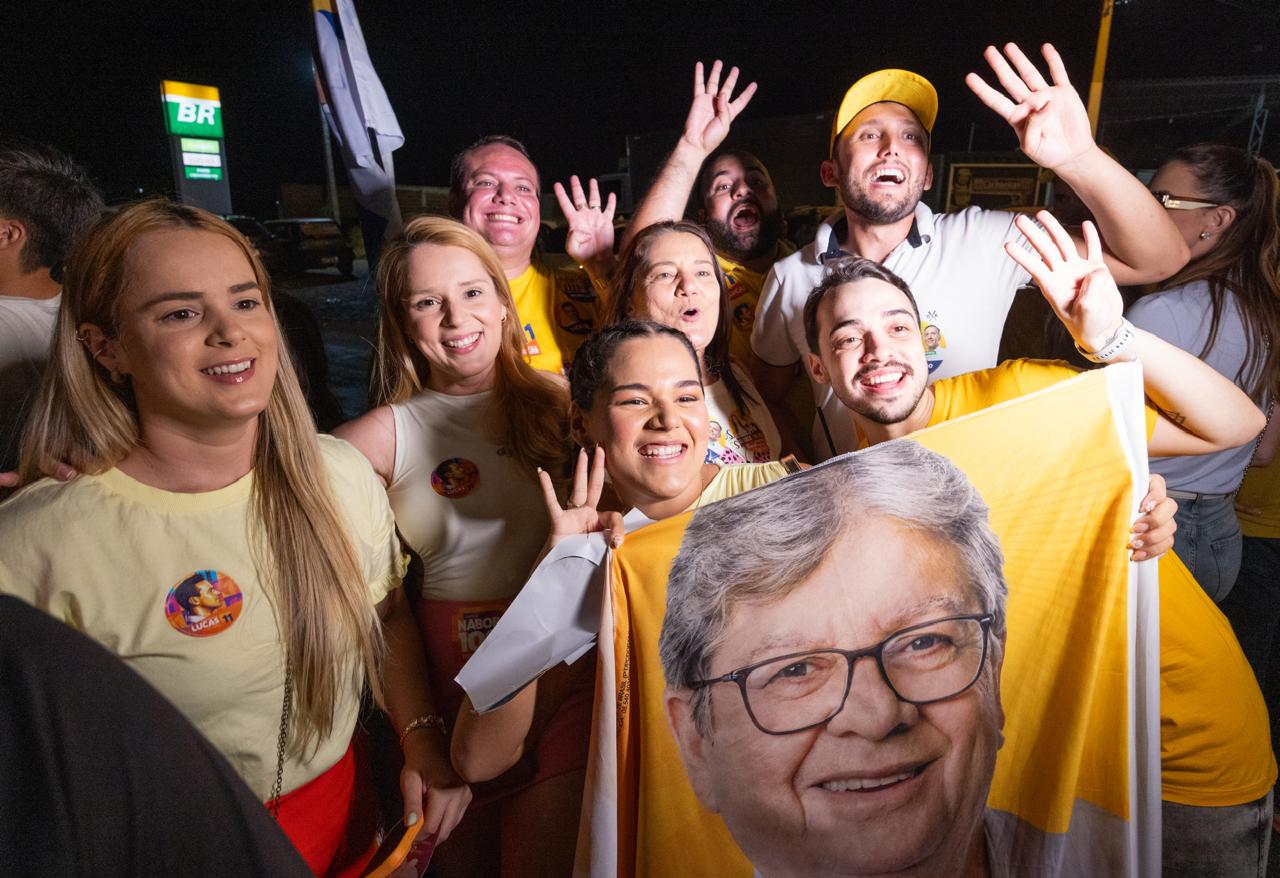 Grupo de jovens apoiadores sorri e acena enquanto segura bandeira amarela estampada com o rosto de João Azevêdo.