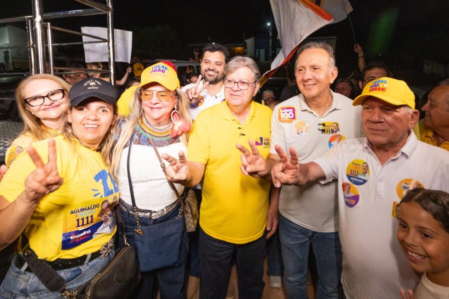 João Azevêdo de camisa amarela posa ao lado do deputado federal Aguinaldo Ribeiro e apoiadores fazendo o número quatro com os dedos.