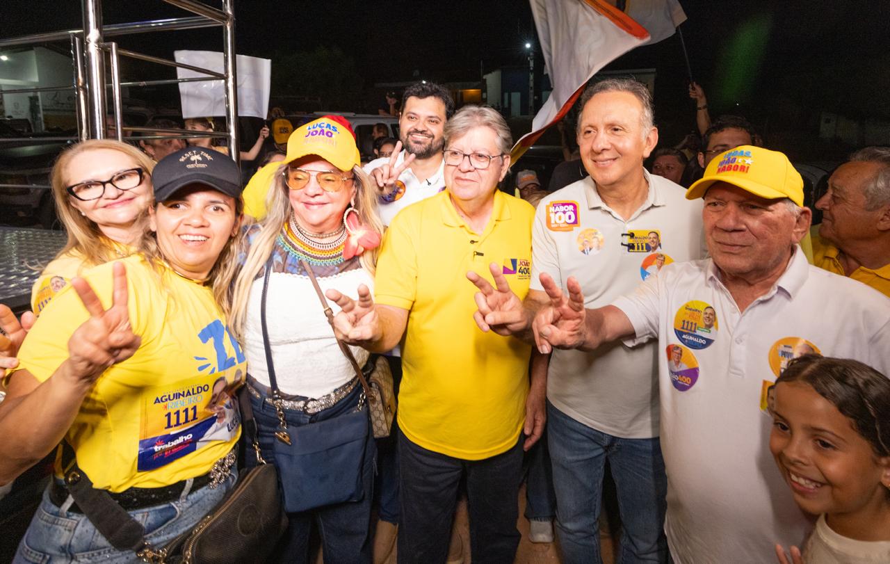 João Azevêdo de camisa amarela posa ao lado do deputado federal Aguinaldo Ribeiro e apoiadores fazendo o número quatro com os dedos.