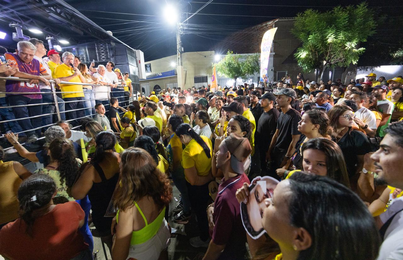 Vista lateral da comitiva no palanque móvel com João Azevêdo discursando e rua tomada por apoiadores à noite.