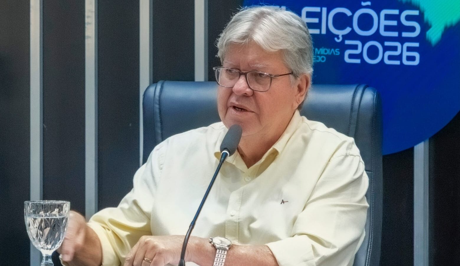 João Azevêdo sentado à mesa de estúdio gesticula em frente ao microfone com painel do programa ao fundo.
