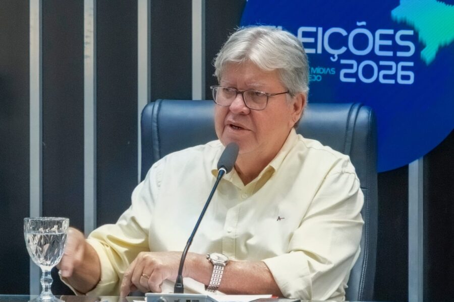João Azevêdo sentado à mesa de estúdio gesticula em frente ao microfone com painel do programa ao fundo.