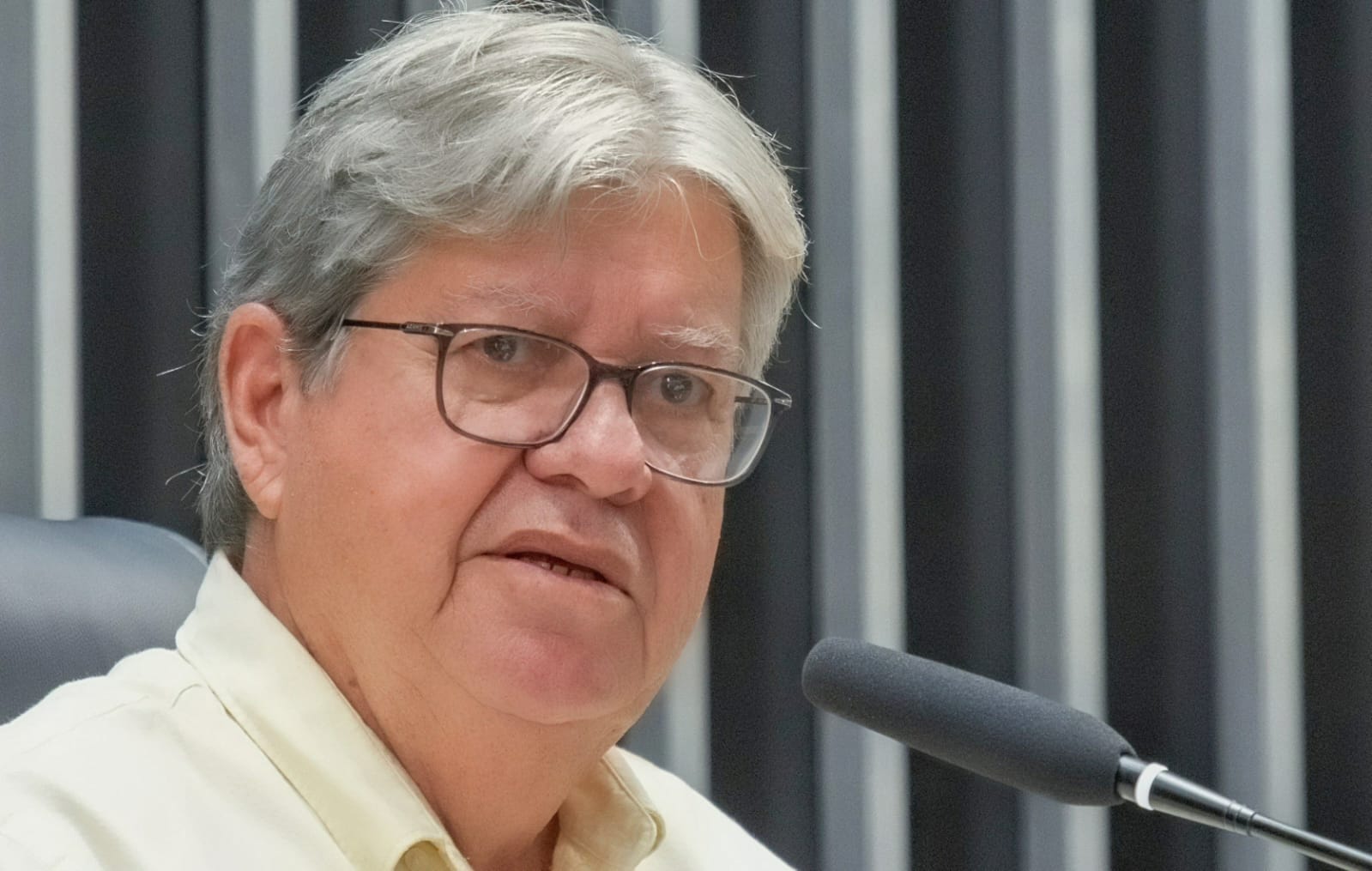 Em close, João Azevêdo fala ao microfone vestindo camisa clara e óculos durante sabatina em estúdio.