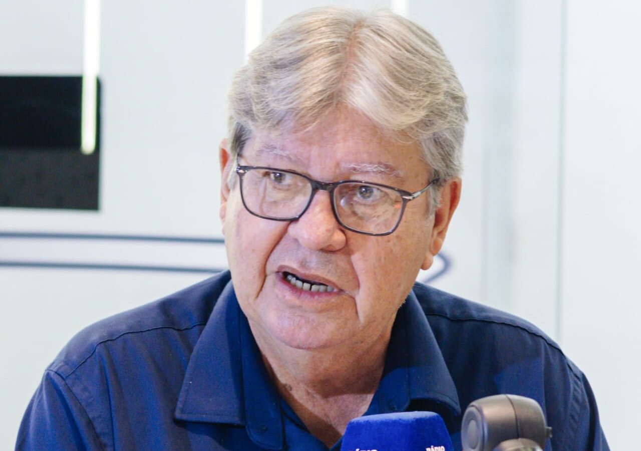 Em close, João Azevêdo veste camisa azul e óculos enquanto fala diante do microfone da BandNews em estúdio de rádio.