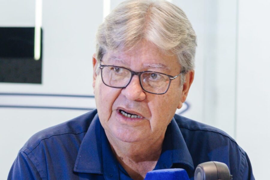 Em close, João Azevêdo veste camisa azul e óculos enquanto fala diante do microfone da BandNews em estúdio de rádio.