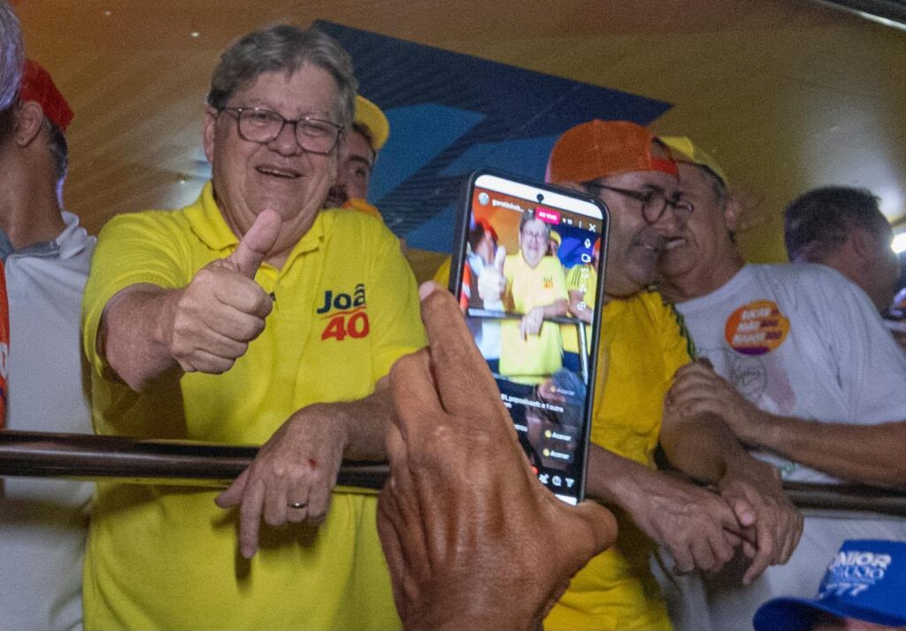 João veste polo amarela "João 40", faz sinal de positivo com o polegar e sorri para a câmera de um celular erguido em primeiro plano.