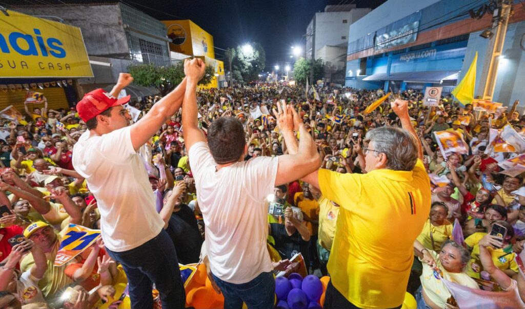 Fábio Tyrone, Lucas Ribeiro e João Azevêdo de costas sobre o trio erguem as mãos unidas para o público presente na rua iluminada.