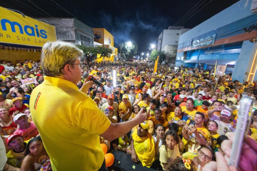 João Azevêdo veste polo amarela e discursa ao microfone do alto da passarela voltado para centenas de apoiadores aglomerados na rua à noite.
