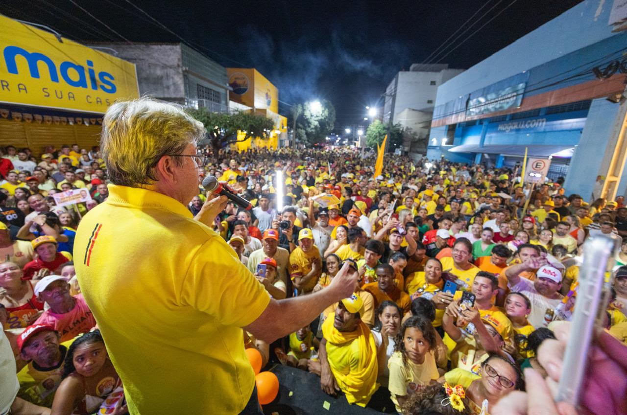 João Azevêdo veste polo amarela e discursa ao microfone do alto da passarela voltado para centenas de apoiadores aglomerados na rua à noite.