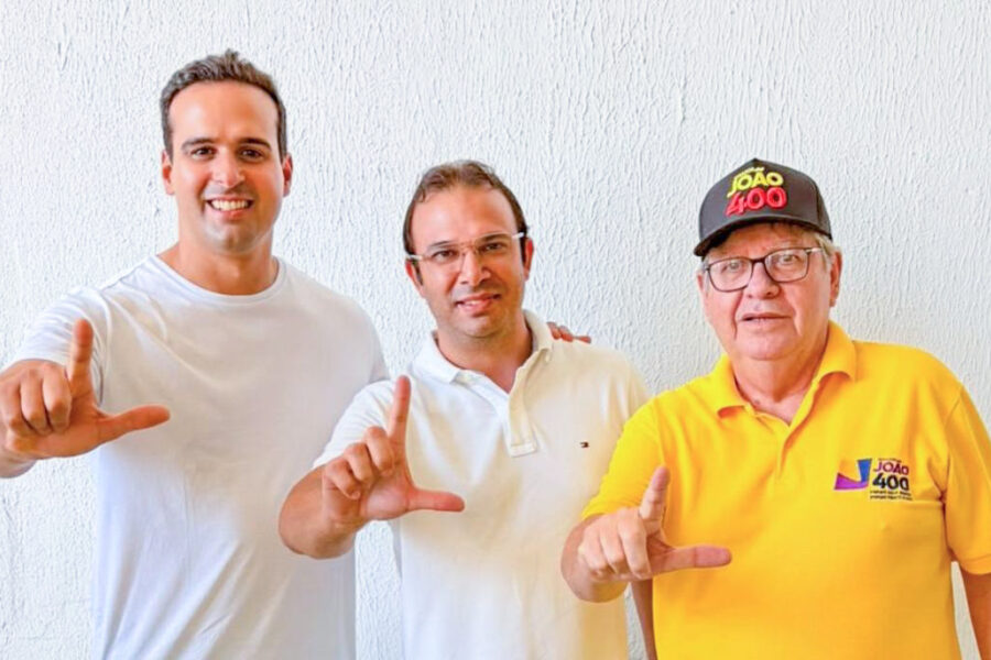 Lucas Ribeiro de camisa branca, Thiago de Claudino ao centro de camisa polo branca e óculos, e João Azevêdo de polo amarela e boné preto posam alinhados fazendo o gesto de "L" com as mãos diante de parede branca clara.