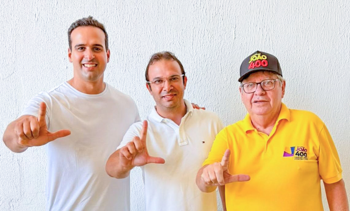 Lucas Ribeiro de camisa branca, Thiago de Claudino ao centro de camisa polo branca e óculos, e João Azevêdo de polo amarela e boné preto posam alinhados fazendo o gesto de "L" com as mãos diante de parede branca clara.