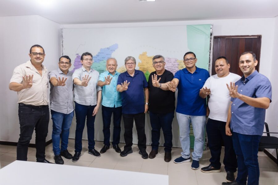 João Azevêdo, o deputado estadual Branco Mendes, o presidente da Câmara Oto Mariano e vereadores de Caaporã posam perfilados em sala fazendo o sinal do número quatro com as mãos espalmadas para a frente.