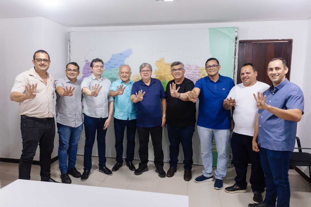 João Azevêdo, o deputado estadual Branco Mendes, o presidente da Câmara Oto Mariano e vereadores de Caaporã posam perfilados em sala fazendo o sinal do número quatro com as mãos espalmadas para a frente.