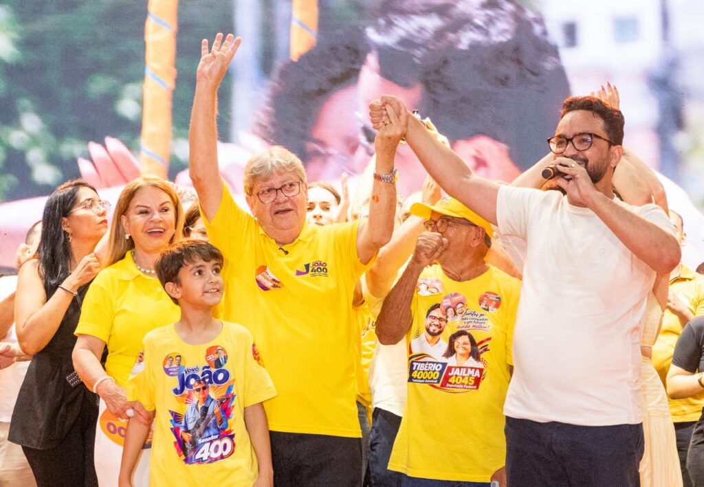 João Azevêdo veste polo amarela e ergue a mão direita enquanto segura a mão de Tibério Limeira no palco, ao lado do neto João Vitor sorridente.