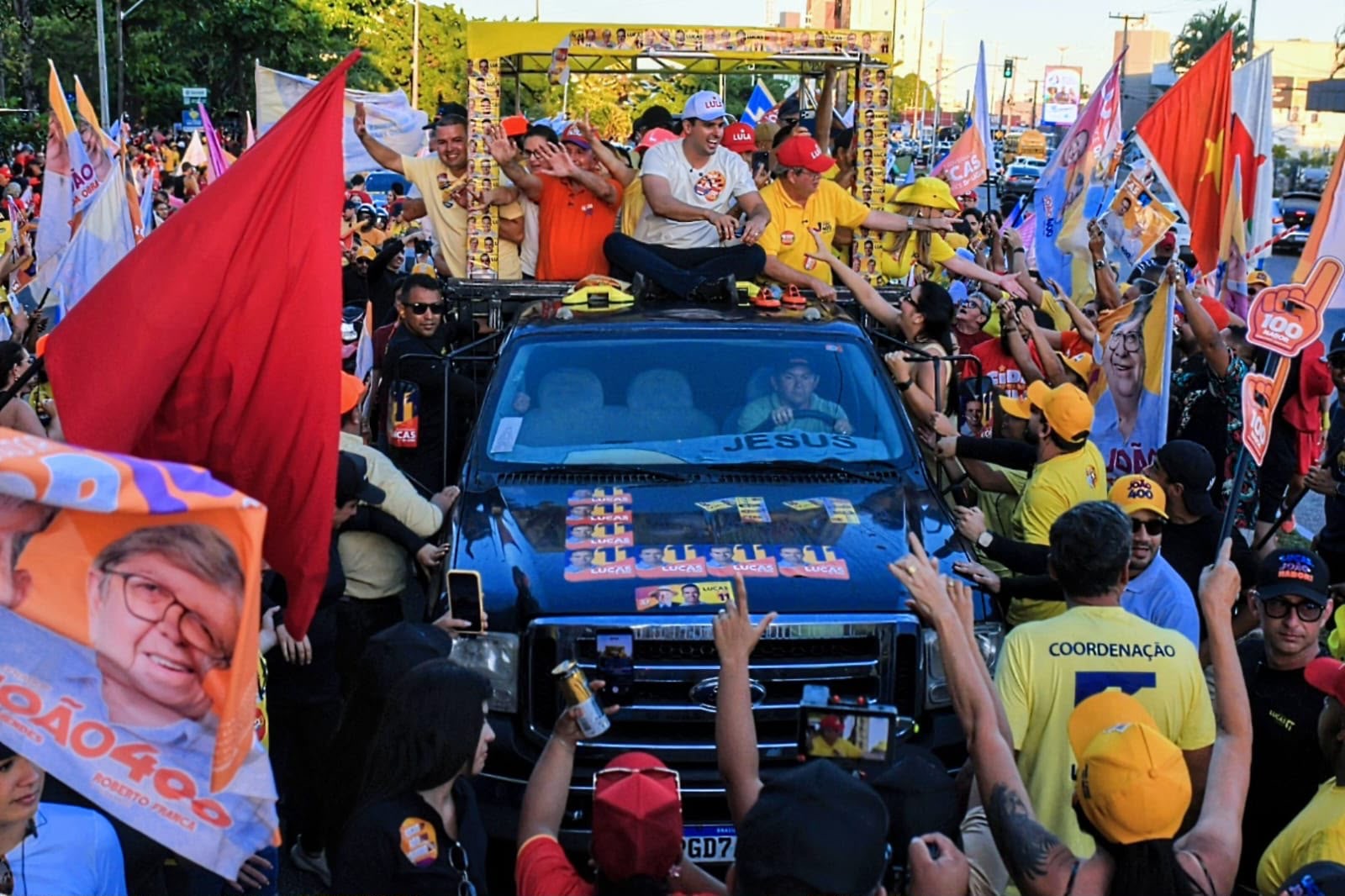 João Azevêdo, Lucas Ribeiro e comitiva em cima de caminhonete cercados por apoiadores com bandeiras coloridas nas ruas de João Pessoa.