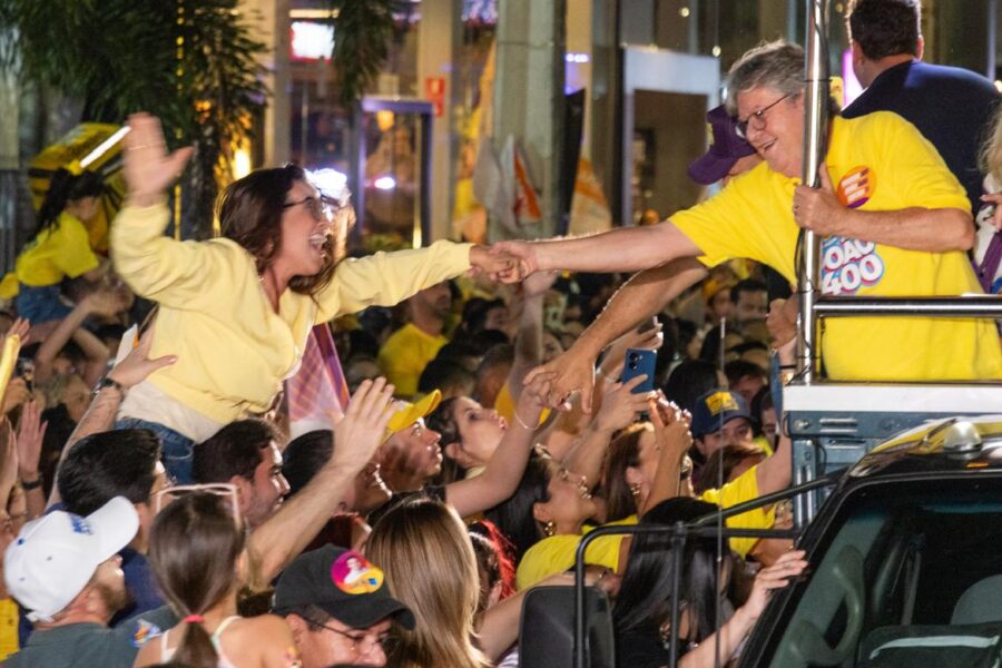 Jovem suspensa por apoiadores veste casaco amarelo e estende a mão com sorriso largo para cumprimentar João Azevêdo no alto do veículo.