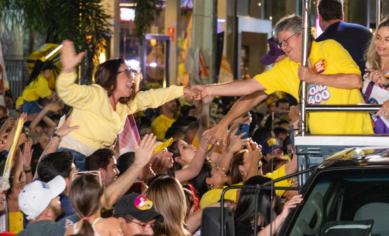 Jovem suspensa por apoiadores veste casaco amarelo e estende a mão com sorriso largo para cumprimentar João Azevêdo no alto do veículo.