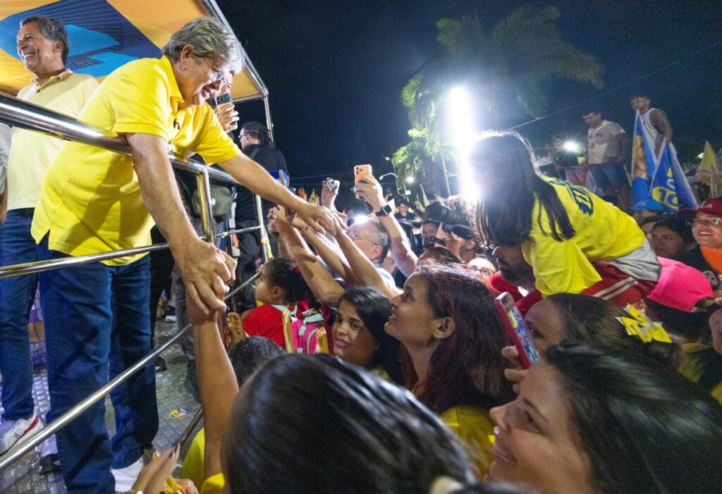 João Azevêdo inclina-se na lateral do veículo e segura a mão de uma mulher e crianças que estendem os braços com entusiasmo para cumprimentá-lo.