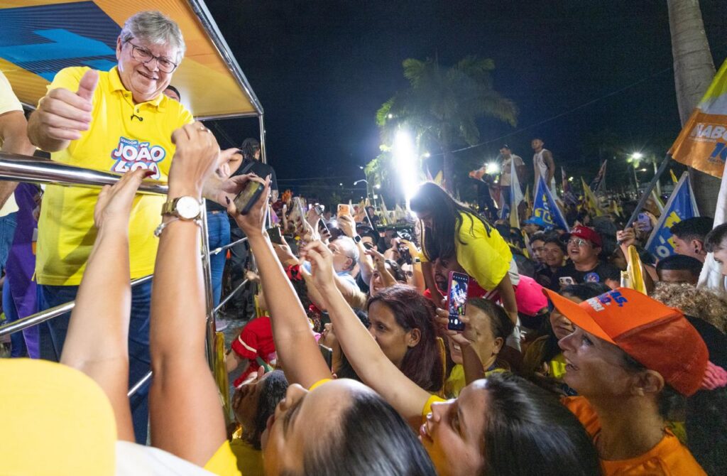 João Azevêdo veste polo amarela de campanha, sorri e faz sinal de positivo com o polegar enquanto apoia a mão no gradil de metal diante de eleitores aglomerados.