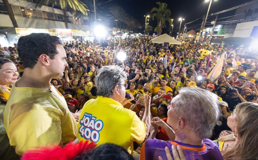 João Azevêdo de costas com polo amarela "João 400" discursa ao microfone ao lado de Lucas Ribeiro voltado para mar de apoiadores em Tambaú.