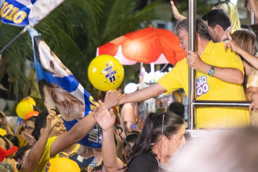 João Azevêdo de polo amarela debruça-se no parapeito do veículo estendendo a mão para eleitores, próximo a balão amarelo com a marca do 11.