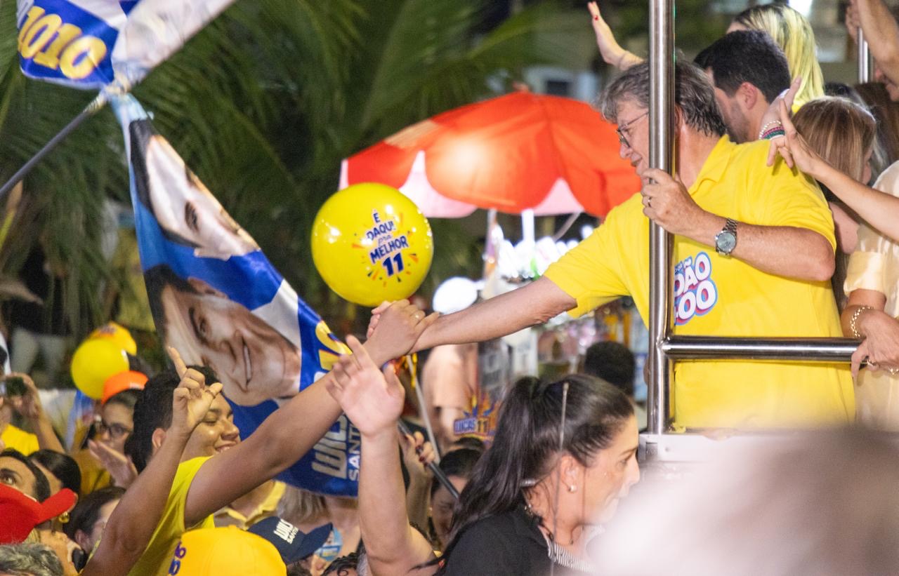 João Azevêdo de polo amarela debruça-se no parapeito do veículo estendendo a mão para eleitores, próximo a balão amarelo com a marca do 11.