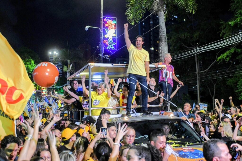 Lucas Ribeiro de pé sobre o teto do carro ergue o punho fechado ao lado de Nabor Wanderley e João Azevêdo diante de letreiro luminoso decorativo.