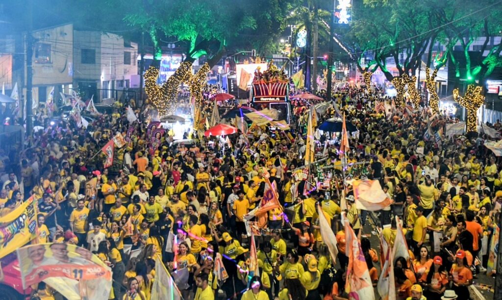 Tomada aérea em plano aberto mostra a Avenida Epitácio Pessoa completamente tomada por milhares de foliões e bandeiras sob iluminação verde nas árvores.